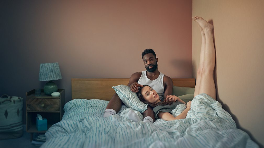 Promotional image for the programme 'Babies' featuring a couple sitting on a bed. The woman is resting her head on a pillow on the man's lap and has her feet and legs above her resting on the wall
