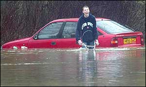 Boy cycles in flooded street in front of submerged car
