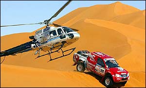 Japanese driver Kenjiro Shinozuka rides over a sand dune as a helicopter monitors his progress