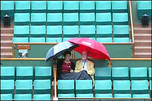 Two spectators huddle under an umbrella as rain delays the start of play