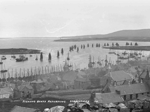 Black and white view over rooftops of Stromness across to fishing fleet returning to harbour.