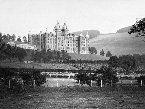 Black and white view of a large, five-storey hotel set on a wooded hillside above fields. The stone building is in Renaissance style, with a symmetrical frontage and a roofline featuring several turrets. A long conservatory extends from one side of the building.