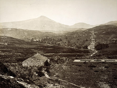 Black and white view showing a small stone shelter and a flat concrete area with mountains behind