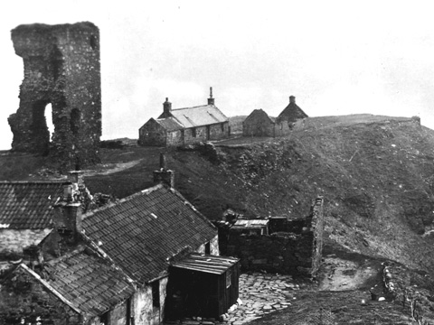 Black and white view of a rocky peninsula on which stand a group of single storey cottages arranged around the ruin of Old Slains Castle.