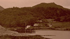 Black and white view across bay to a wooden pier with scattered cottages and hills behind. Two boats lie pulled ashore in the foreground..