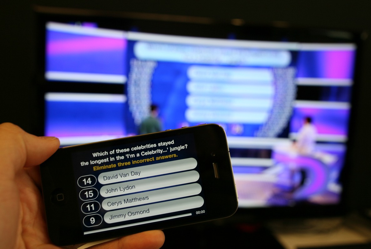 A mobile phone showing a question screen from the Secret Fortune companion application. In the background, a television showing the programme can be seen.
