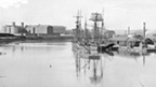 Black and white view of Perth Harbour from the south. A tall sailing ship is docked at the harbour which protrudes in to the river.
