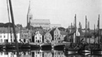 Black and white view of harbour filled with sailed fishing boats with town dominated by church tower in the background