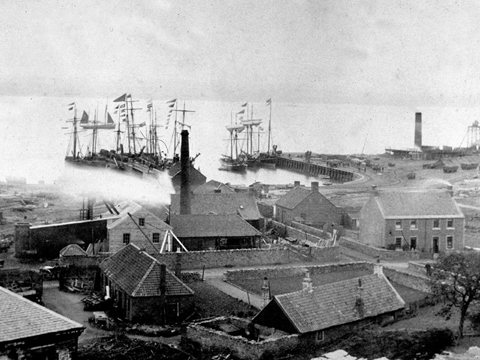 Black and white view over rooftops and industrial chimneys to sailing boats in harbour.
