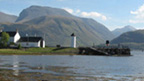 Colour view from a rocky beach across Loch Linnhe to Ben Nevis. In the middle distance is a quayside featuring a cylindrical tower, pitched-roofed buildings and a wooden pier with slipway.