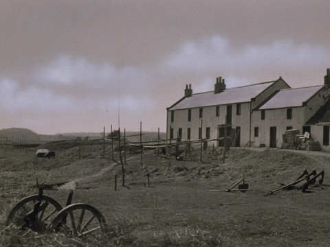 Black and white view of row of stone buildings with wheels, anchors and drying poles in the grass hollow below. A hill and bridges are seen in the in the distance.