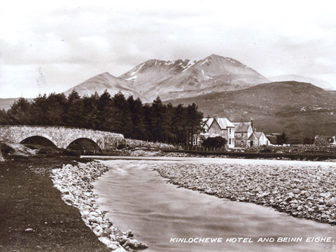 Black and white view of a braided river crossed by a twin-arched road bridge. Behind stands the Kinlochewe Hotel with the mountain Sgurr Ban in the background.