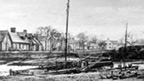 Black and white view of sailing boat in small harbour with row of cottages behind.