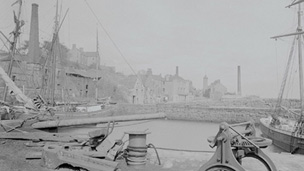 Black and white view of a harbour in which three tall-masted sailing ships are docked. In the foreground are a winch and scattered pieces of dockyard equipment. A village featuring traditional stone buildings, a tollbooth and several tall, industrial chimney stacks, rises up a hill behind the harbour.