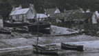 Black and white view showing small and large fishing boats pulled up onto the shore with cottages beneath tall cliffs behind.