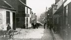 Black and white view down New Street, Paisley. The cobbled street is lined by one and two-storey buildings. A number of people in Edwardian dress, including a man resting against a two-wheeled cart, look towards camera.