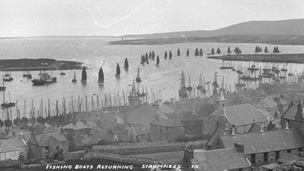 Black and white view over rooftops of Stromness across to fishing fleet returning to harbour.