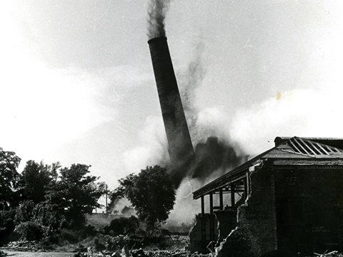 Black and white view of the demolition of a tall, industrial chimney stack.