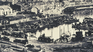 Black and white view of harbour with fleet of fishing boats and a number of loaded trucks on the quayside.