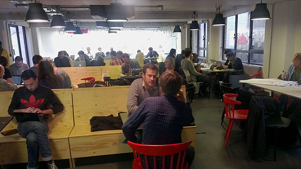 Dozens of people working on wooden benches and red chairs in a brightly lit room