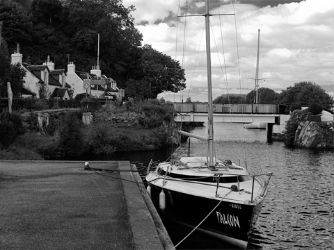 A small yacht, moored at the canal-side, with a bridge over the canal and a row of stone cottages behind.