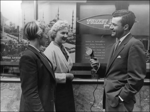 BBC Türkçe yapımcısı Özcan Ergüder, Piccadilly semtindeki bir seyahat acentasının camında asılı İstanbul resimlerini hayranlıkla inceleyen iki İngiliz kızla mülakat yapıyor. İki Şehir programının o ayki bölümü Londra'nın Batısı ve İstanbul'un Avrupa yakasını buluşturuyordu. 14/06/1956 © BBC
