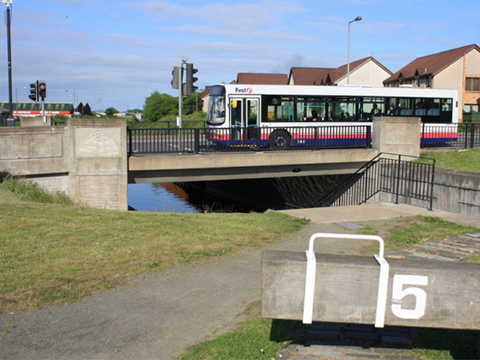 Colour view from tow-path showing a bus crossing a bridge over the canal. Part of a lock-gate, labelled number five, can be seen in the foreground.