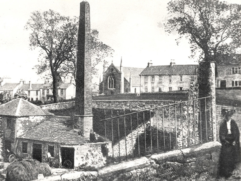 A woman in Edwardian dress stands next to a small set of mill buildings featuring a tall chimney stack. Behind is a row of dwellings and a church