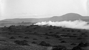 A figure tends to a smoking fire in a field covered with small heaps of seaweed.