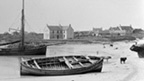 Black and white view of Scarinish, showing a curving, sandy bay in which an open-decked, single-masted boat and a smaller dinghy are moored. Another open decked boat lies beached in the foreground. A number of cows can be seen on the beach and there is a scattered collection of single and two-storey buildings in the background.