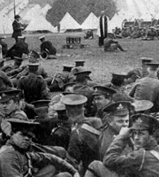 Black-and-white photo from 1915. Soldiers in First World War uniform sit on the ground, some looking at the camera, while a Church of England clergyman speaks. In the background are rows of tents