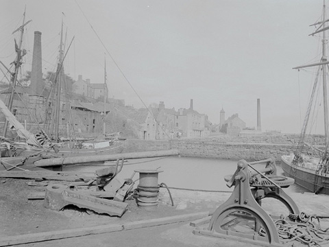 Black and white view of a harbour in which three tall-masted sailing ships are docked. In the foreground are a winch and scattered pieces of dockyard equipment. A village featuring traditional stone buildings, a tollbooth and several tall, industrial chimney stacks, rises up a hill behind the harbour.