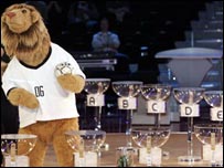 Mascote ao lado dos recipientes em que será feito o sorteio dos grupos da Copa