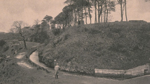 A small figure stands on a footpath by a narrow man-made water course at the foot of a hill with trees around.