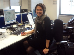 Picture of Catherine Carr at her desk at work wearing a black dress with black tights and boots.