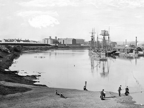 Black and white view of Perth Harbour from the south. A tall sailing ship is docked at the harbour which protrudes in to the river. In the foreground, a group of men are fishing while another man in a top hat sits sketching or painting the scene. In the background are some large, possibly industrial buildings.