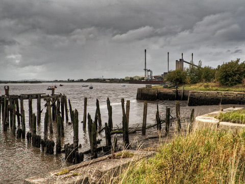 Colour view of derelict harbour at Alloa. In the foreground are the remains of a timber pier, with a stone harbour wall visible on the other side of a wet dock. Several industrial chimneys can be seen beyond this.