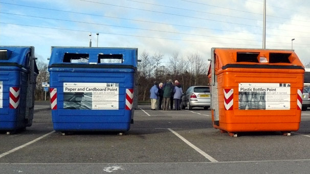 People in a car park, between recycling bins