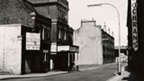 Black and white view along deserted street. A dilapidated theatre building to the left features a hoarding advertising the boy scouts of paisley gangshow of 1959. A raised railway line can be seen at the end of the street.