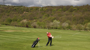 A single golfer stands mid swing on a sunny golf course with tall, flat topped hills behind under a threatening sky of dark clouds.