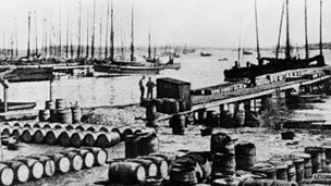 Black and white view of Baltasound Harbour. The quayside is covered with many rows of barrels. A number of single and two-masted boats are moored in the background.