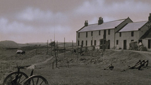 Black and white view of row of stone buildings with wheels, anchors and drying poles in the grass hollow below. A hill and bridges are seen in the in the distance.
