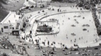 Black and white view, looking down from coastal cliffs to twin open air pools, with an Art Deco pavilion to the left of frame. One pool contains a mix of people paddling and rowing boats. The pool to the right is surrounded by a crowd of people, possibly watching a swimming competition. A large number of people are sunbathing on the surrounding grassy area.