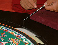 A monk holds the chak-pur rod, a pen-sized metal implement ridged along its length, and scrapes it with a smaller tool to make the sand flow freely from its end
