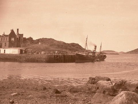 Black and white view of a fishing boat berthed at a simple harbour. The burned out shell of a large house stands in the background.