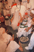 Man sitting on chair with his head covered by silver fringe all the way to the floor. Female worshippers prostrate before him