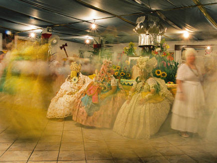 Three women seated on chairs in a large hall. Wearing masks, other dancers swirl around them