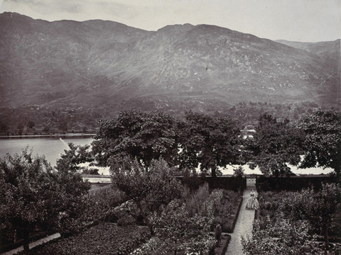 Black and white view of a formally laid out garden with trees and possibly vegetable beds. Between the beds are parallel paths leading down to a loch with hills behind. A woman in Victorian dress stands on one of the paths.