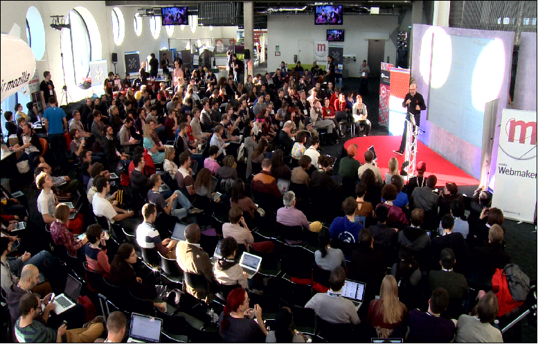 The main arena at Mozfest 2012 in London, Greenwich. Picture shows a large room in the Ravensbourne building with a speaker on a platform. He is being watched by hundreds of people.