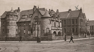 Black and white view of two-storey hospital building featuring a range of pitched-roofed main blocks with turrets. The hospital stands at the crossroads of two wide streets along which run a number of tram tracks.